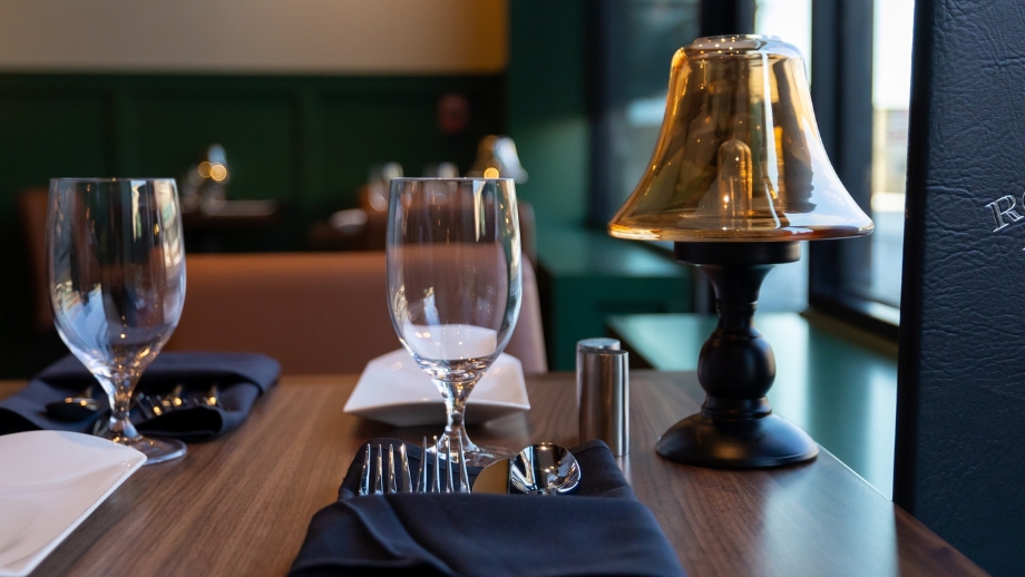 A table set for two in a restaurant with a wooden table, black napkins, wine glasses, and a brass lamp.