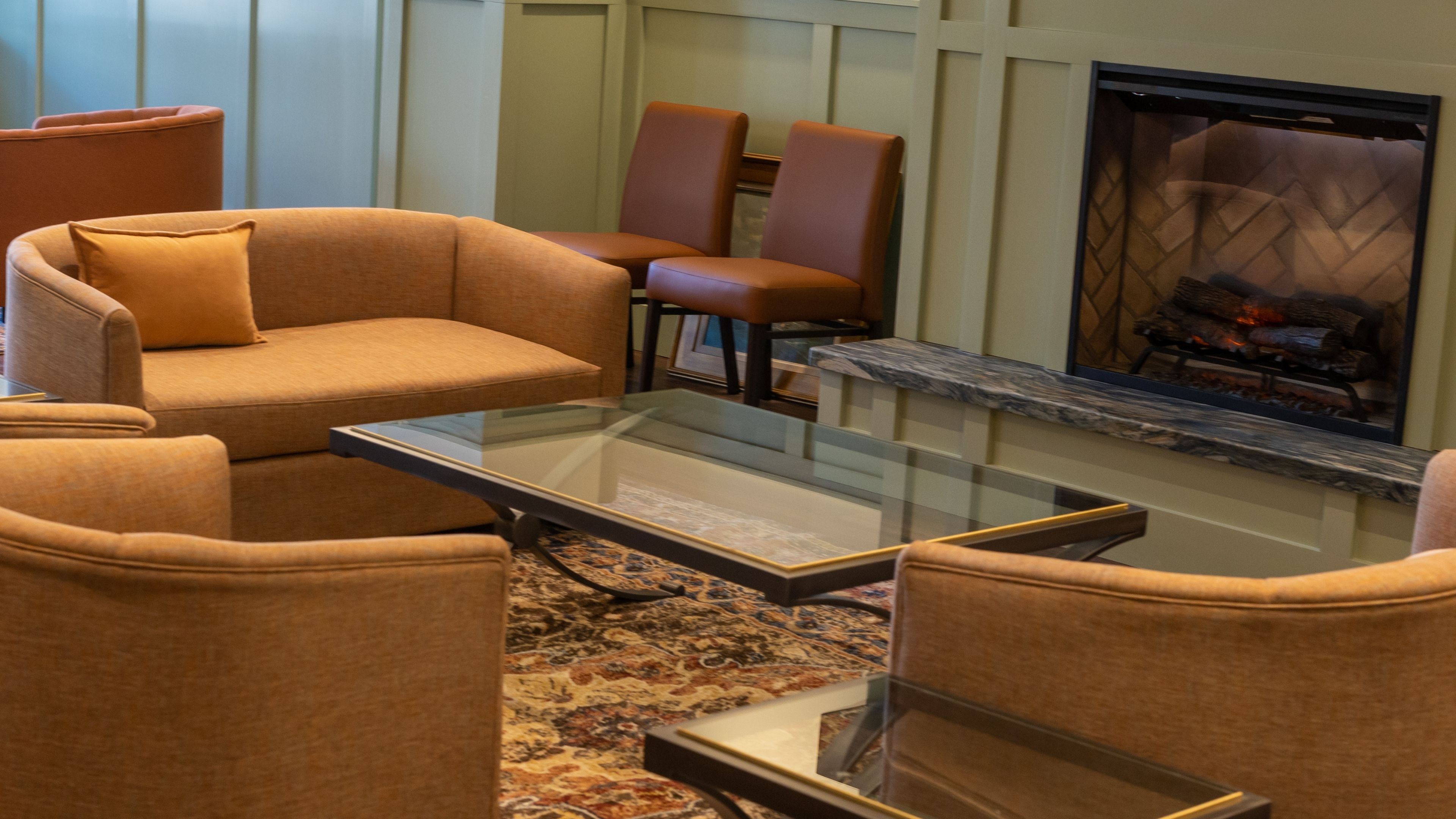 A cozy hotel lobby seating area with a fireplace, comfortable chairs and sofa, and a glass coffee table.