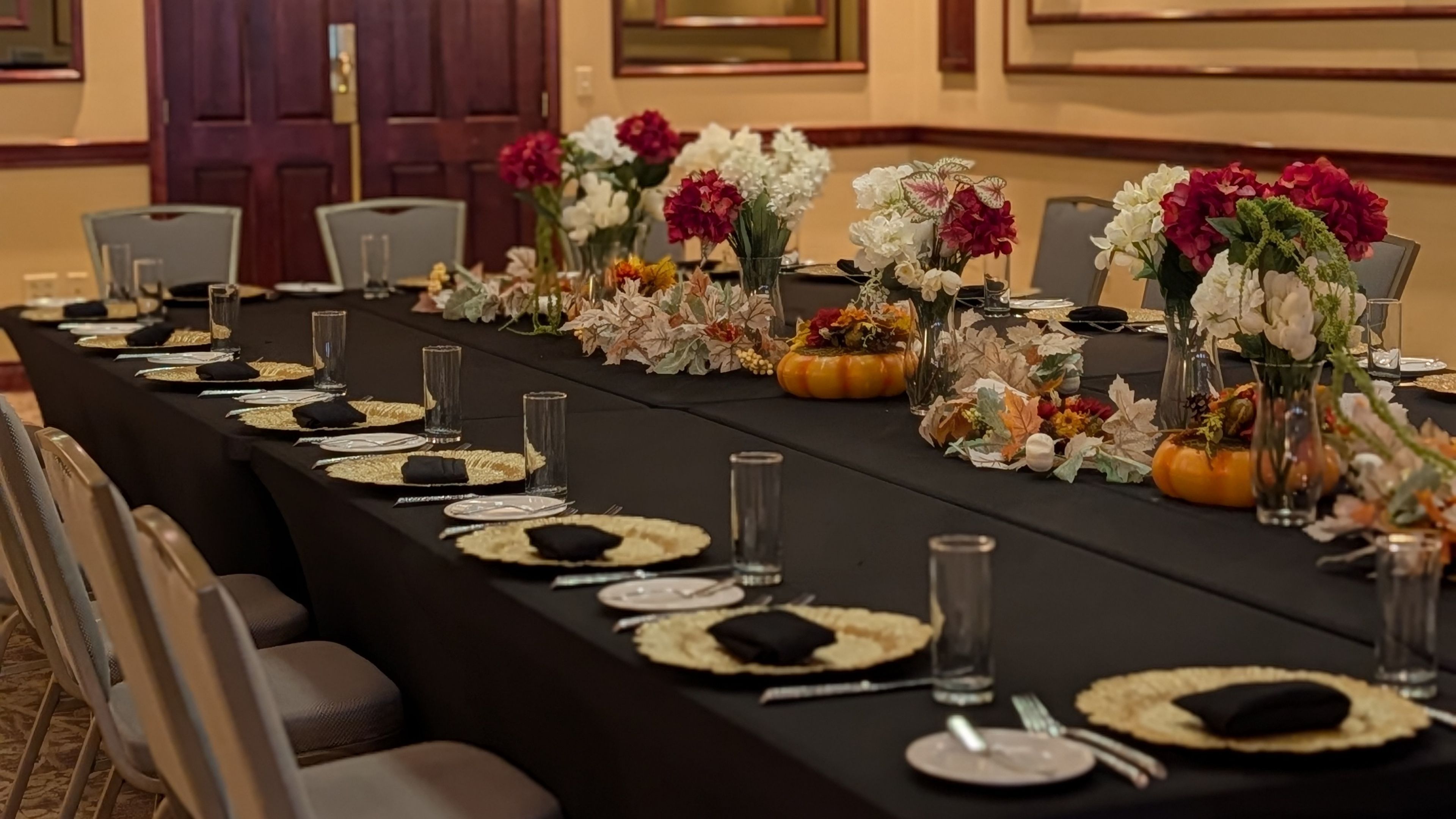 A long table set for a formal meal with a black tablecloth, flowers, pumpkins, and fine china.
