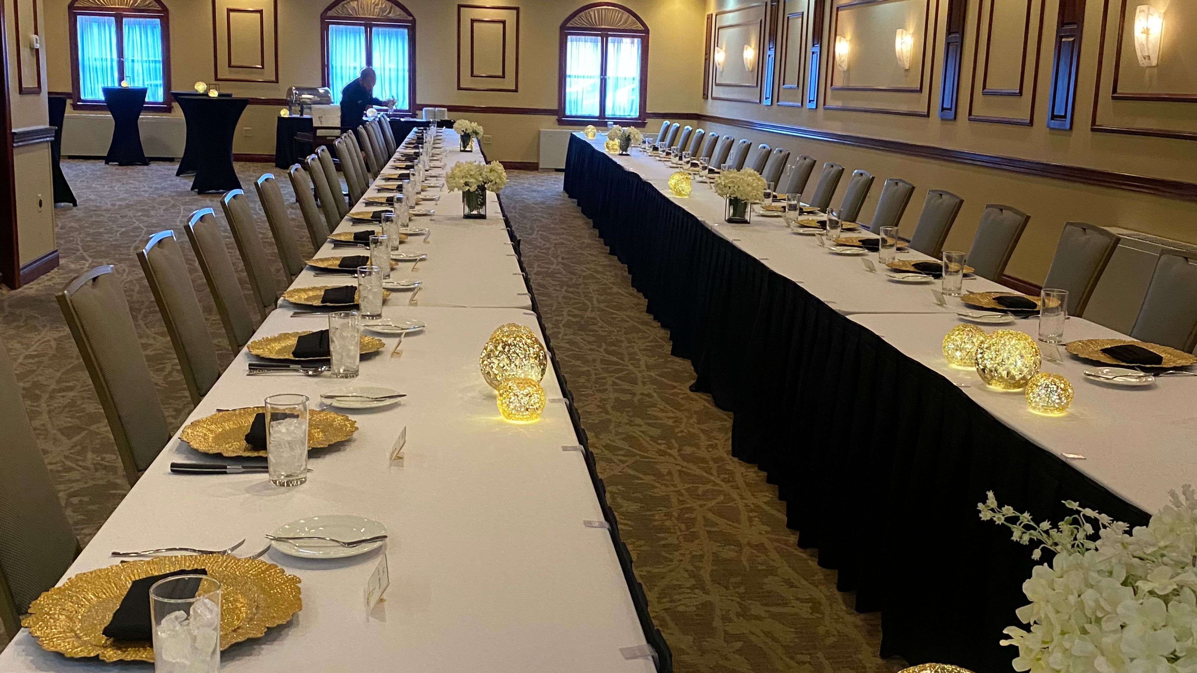 A long, formal dining table set with white tablecloths, gold plates, black napkins, and glassware.