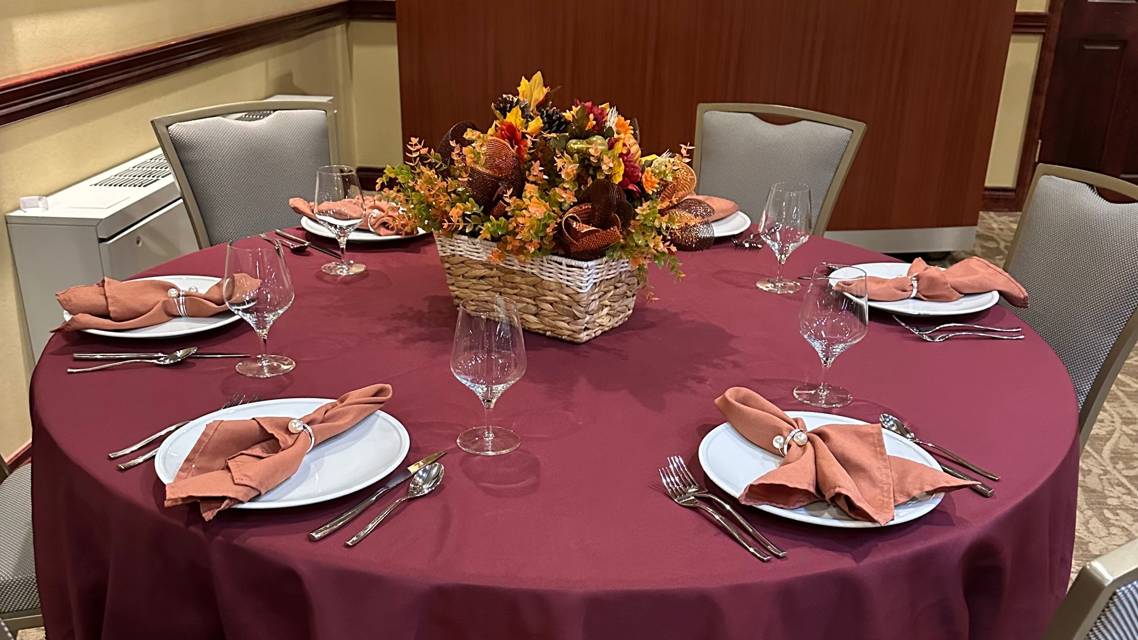 A round dining table set with a maroon tablecloth, white plates, orange napkins, wine glasses, and silverware, with a floral centerpiece.