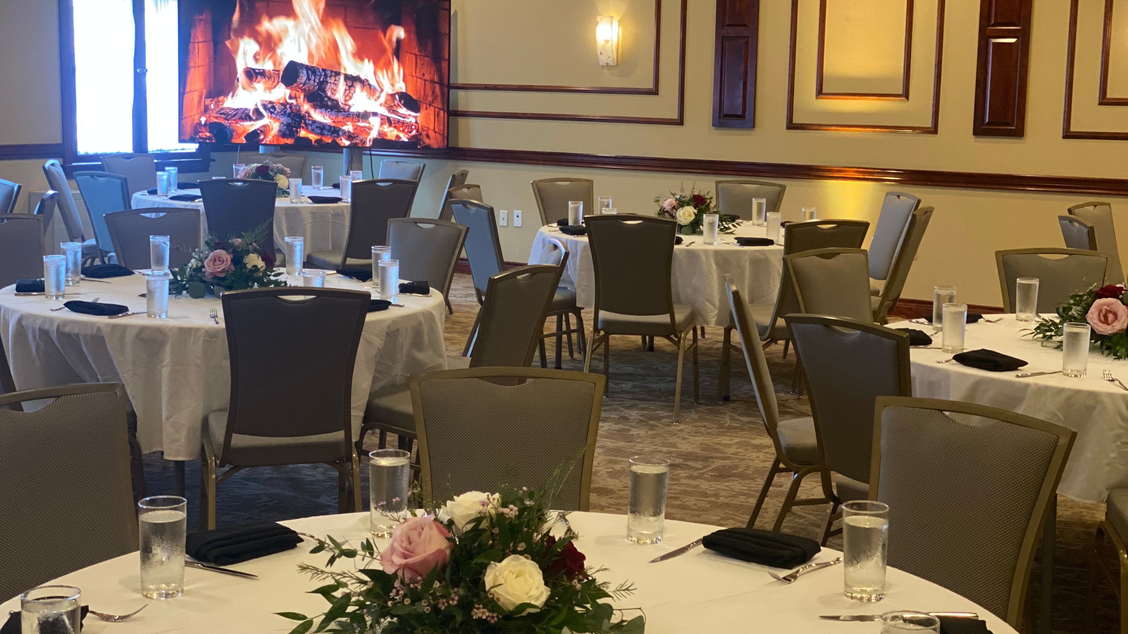 A well-set dining room with round tables, white tablecloths, flower arrangements, and a fireplace screen on the wall.